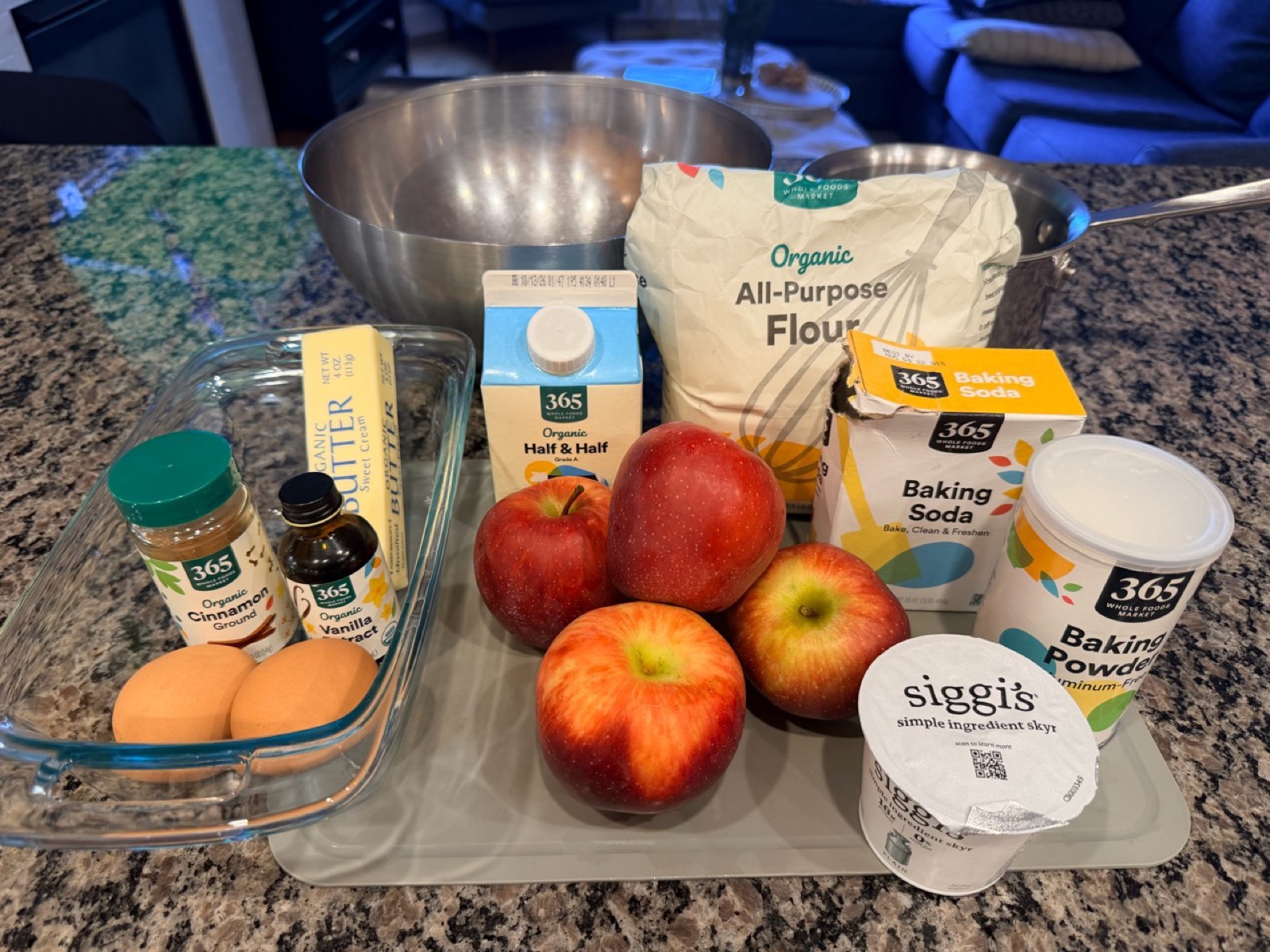 Apples, flour, butter, eggs, cinnamon, vanilla, yogurt, half-and-half, and leaveners arranged on the kitchen counter.