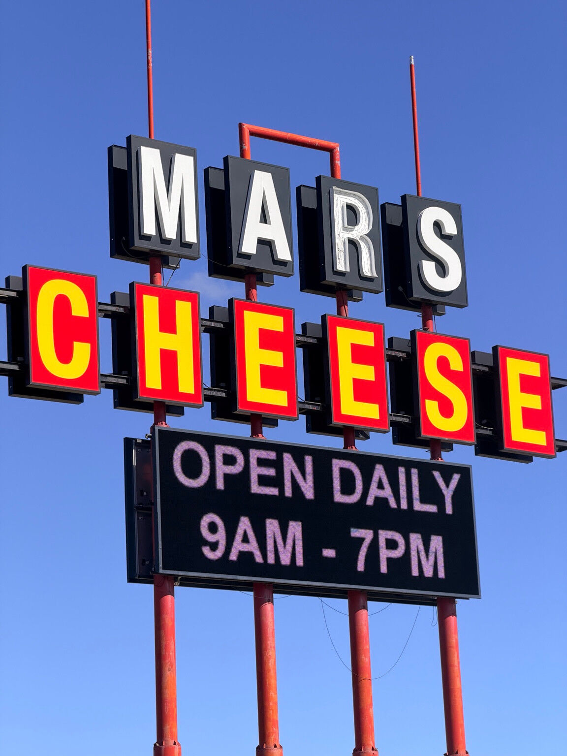 The Mars Cheese sign against a blue sky, with a digital panel showing daily opening hours.